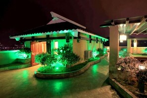 Una casa pequeña con techo inclinado, rodeada de un jardín con iluminación verde y una zona de piscina con pérgola.