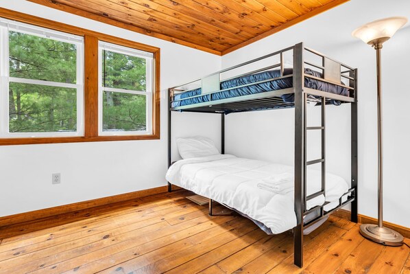 Habitación con literas, suelo de madera, ventana con vistas a árboles y una lámpara de pie.