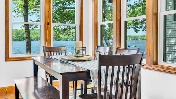 Un comedor con una mesa y sillas, ventanas amplias con vista a árboles y agua, y una vela sobre la mesa.