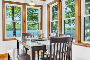 Un comedor con una mesa y sillas, ventanas amplias con vista a árboles y agua, y una vela sobre la mesa.