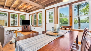 Una habitación amplia con techo de madera, ventanas grandes, una mesa de comedor con sillas, un sofá y un televisor sobre un mueble.