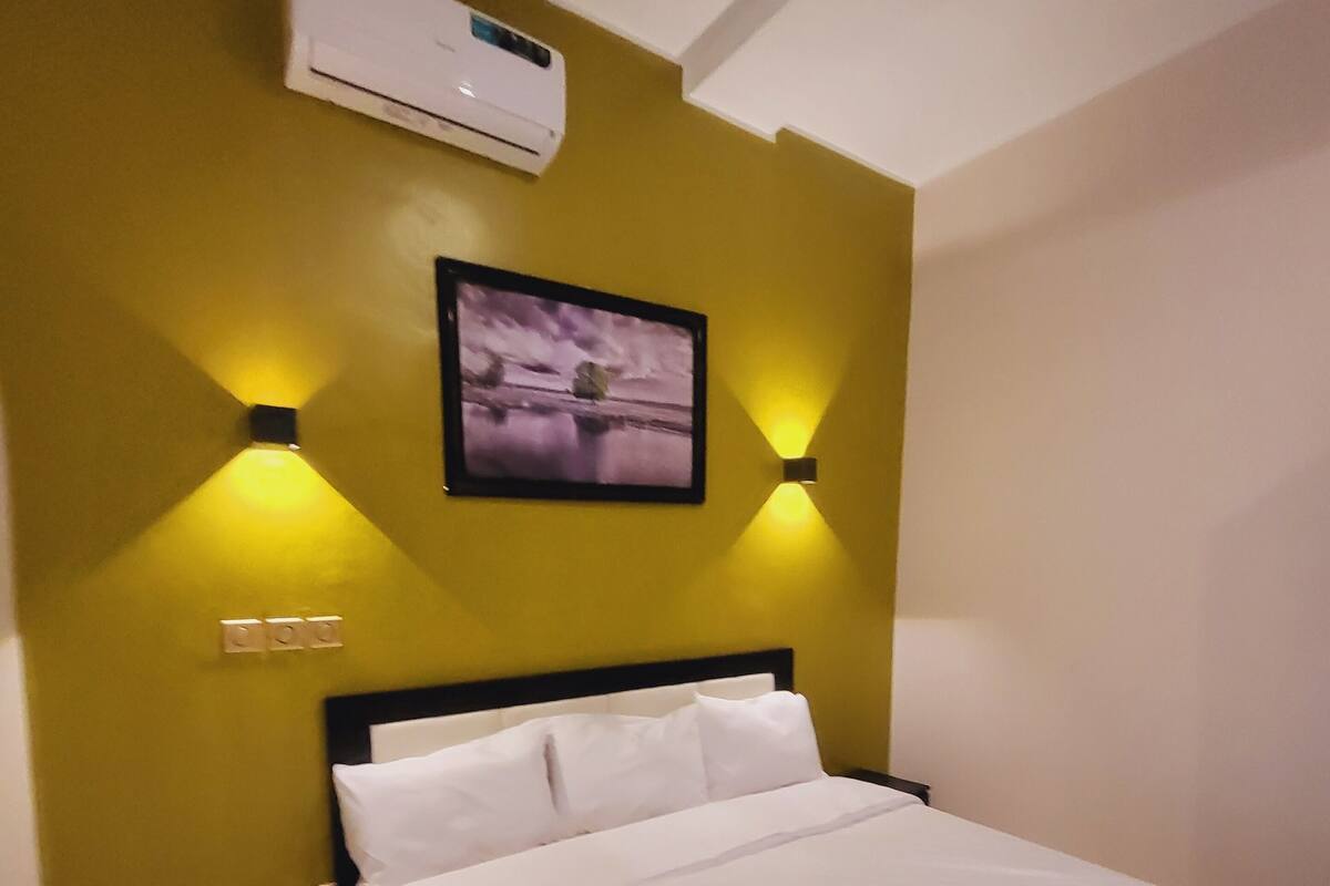 Een hotelkamer met een bed, een airconditioning, twee wandlampen en een ingelijst schilderij aan de muur.
