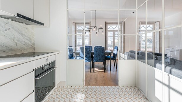 Cocina moderna con armarios blancos y encimera de mármol, que da paso a un comedor con sillas tapizadas en azul y suelo de madera.