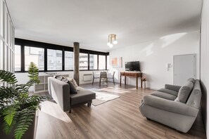 Sala de estar moderna com sofá cinzento, mesa de centro de madeira e televisão sobre suporte de madeira. Grandes janelas oferecem luz natural e vista para a cidade.