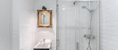Ein modernes Badezimmer mit weißen Fliesen, einer Duschkabine aus Glas, einem schwarzen Mülleimer und einem spiegel mit goldener Rahmen.