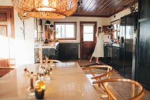 A kitchen with a dining area, wooden cabinets, a large window, and a hanging light fixture. - Moon Shadow Ranch ★  Desert Medicine Retreat (Twentynine Palms)