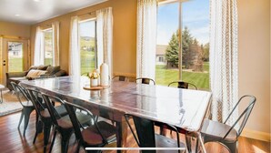 Ein Essbereich mit großem Tisch und Stühlen, einer Couch und Blick auf eine grüne Landschaft durch große Fenster.