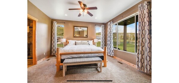 A bedroom with a large bed, a ceiling fan, a window with patterned curtains, and a view of the outdoors.