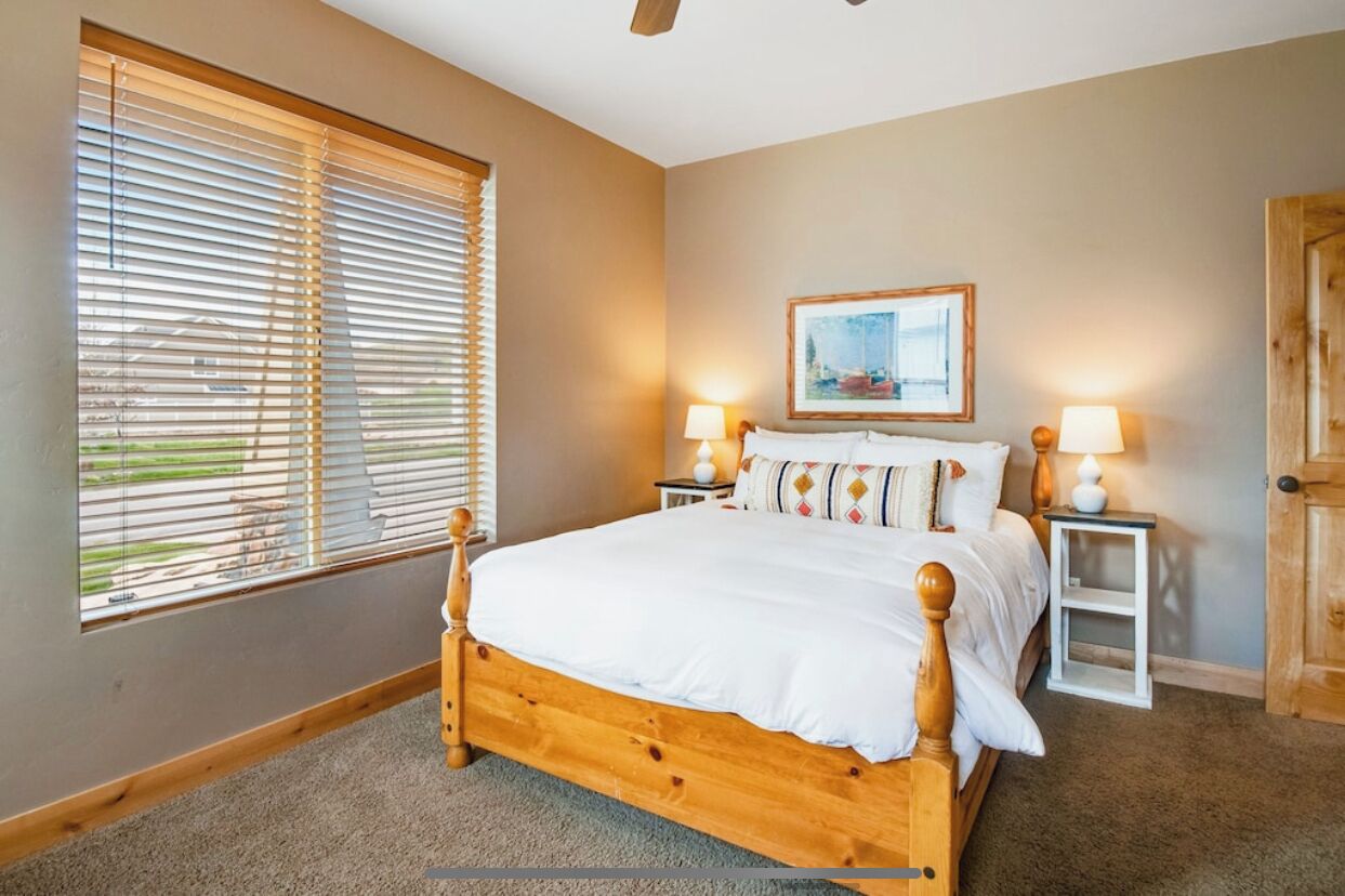 A bedroom with a wooden bed, two bedside tables with lamps, a window with blinds, and a door.