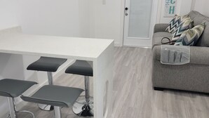 En kompakt opholdsstue med et hvidt tekøkken, en grå sofa med mønstrede puder, og en gennemsigtig plastikpose på sofaen.