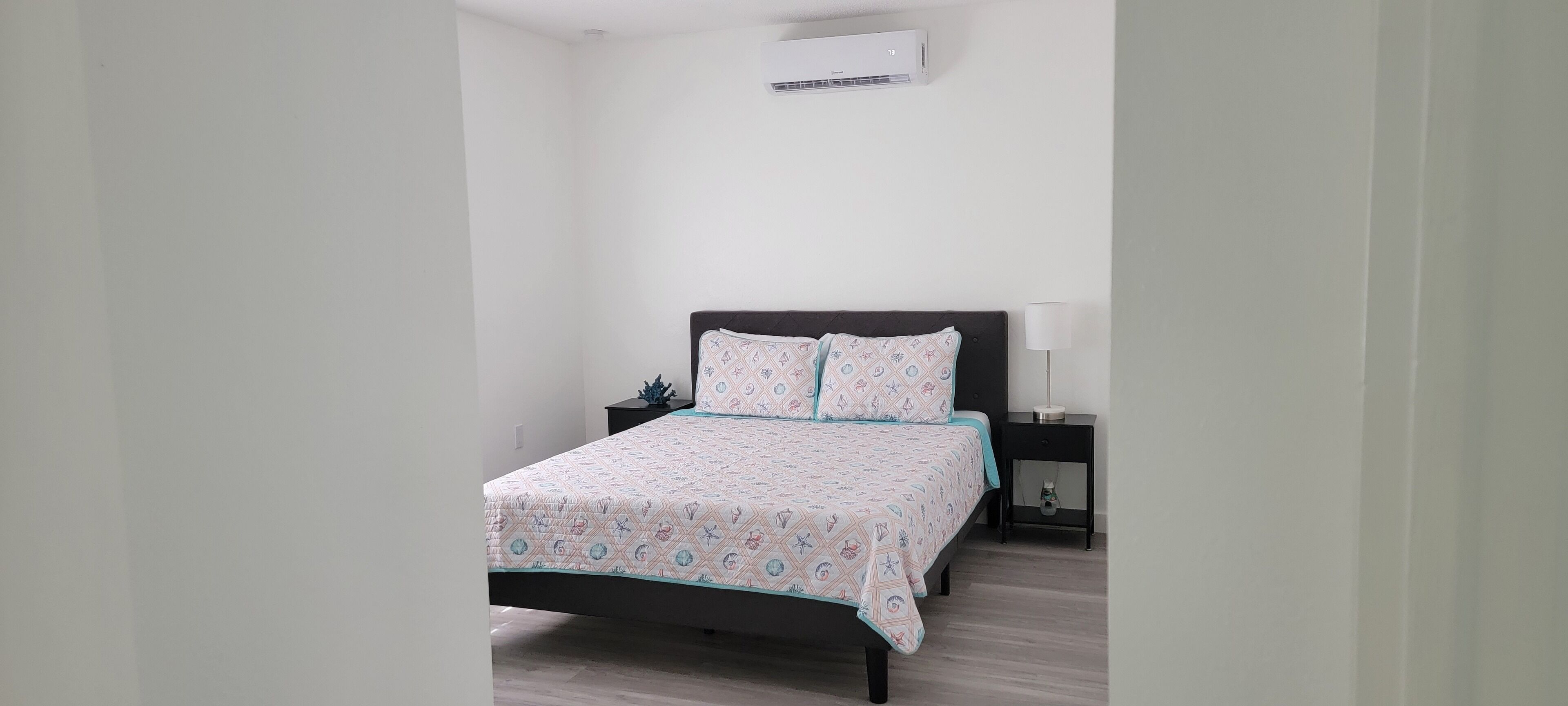 A bedroom with a bed, a nightstand, a lamp, and an air conditioning unit on the wall.