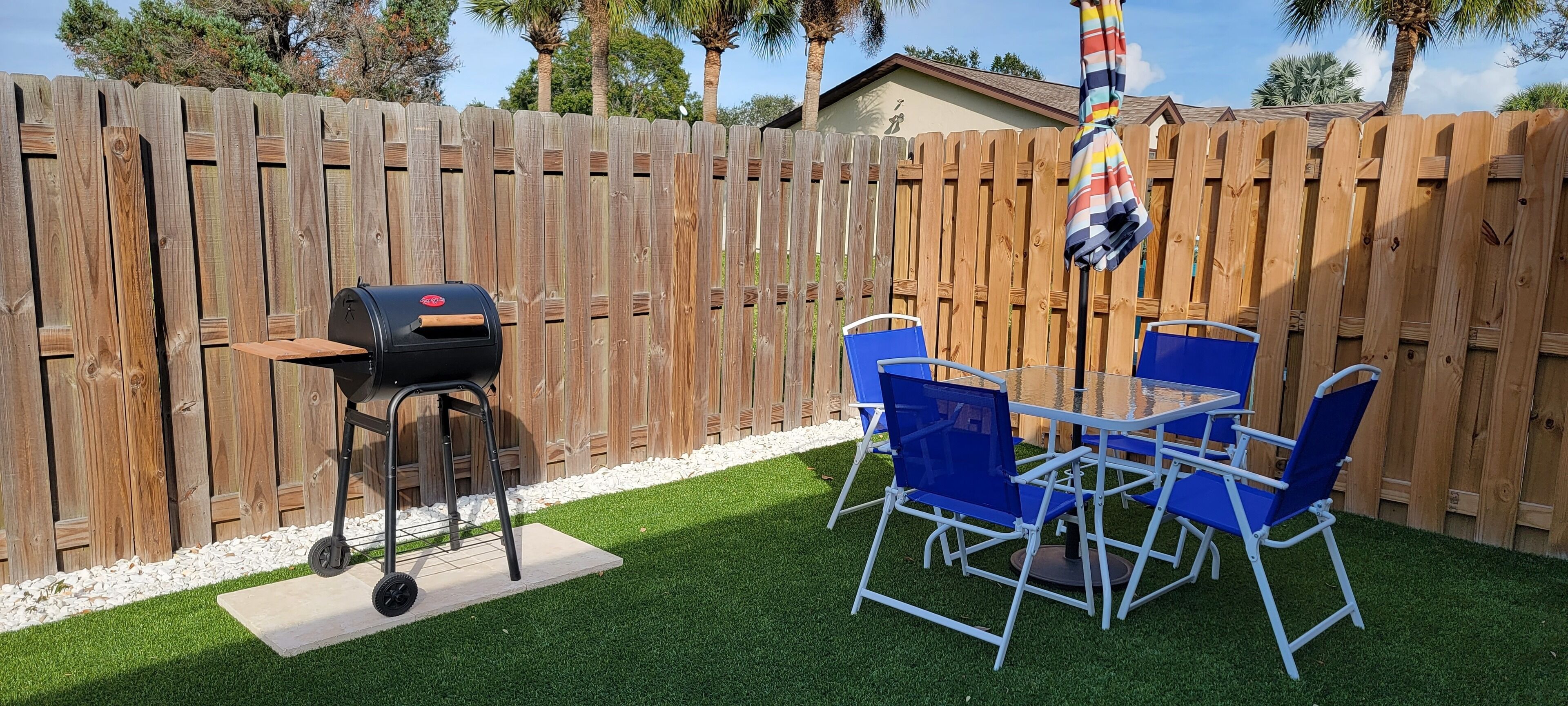 A backyard with a wooden fence, a barbecue grill, and blue chairs around a table.