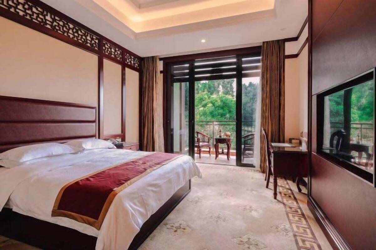 Una habitación de hotel con cama, escritorio, televisor y balcón con vistas a la vegetación.