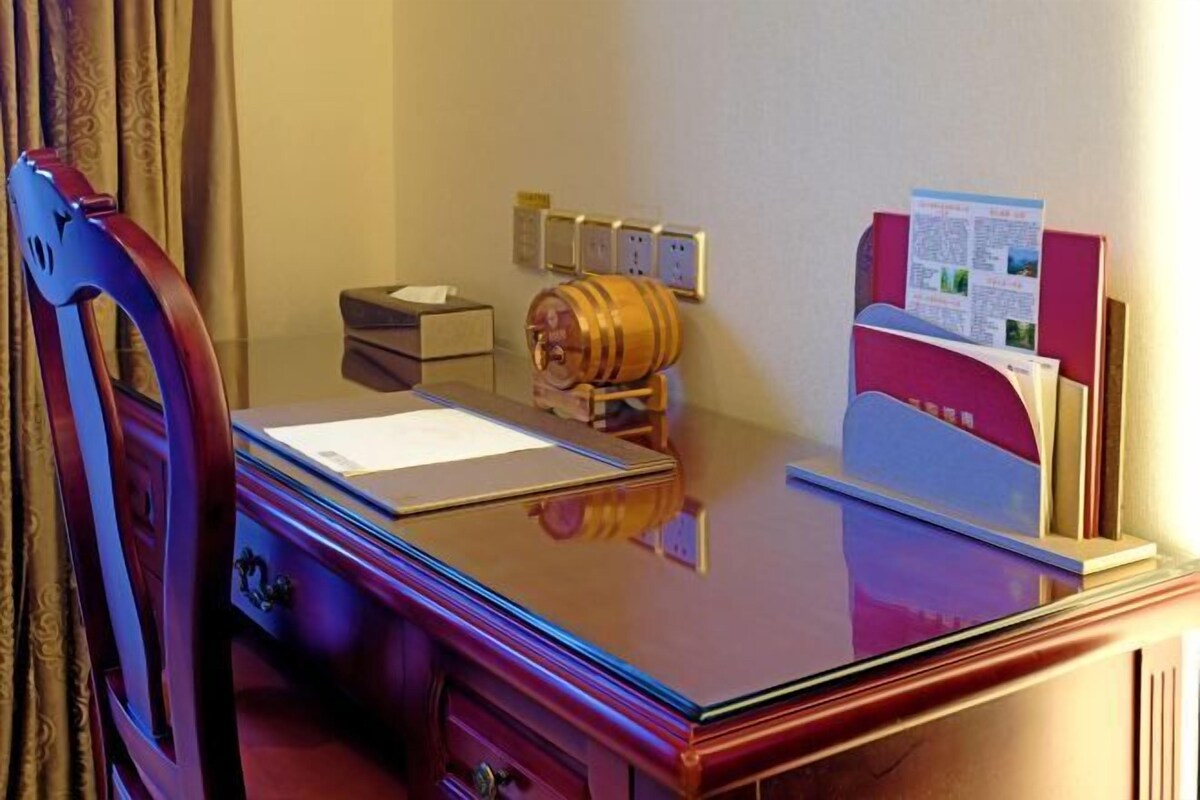 A hotel room with a wooden desk, a chair, a minibar, a small safe, and a bulletin board with papers.