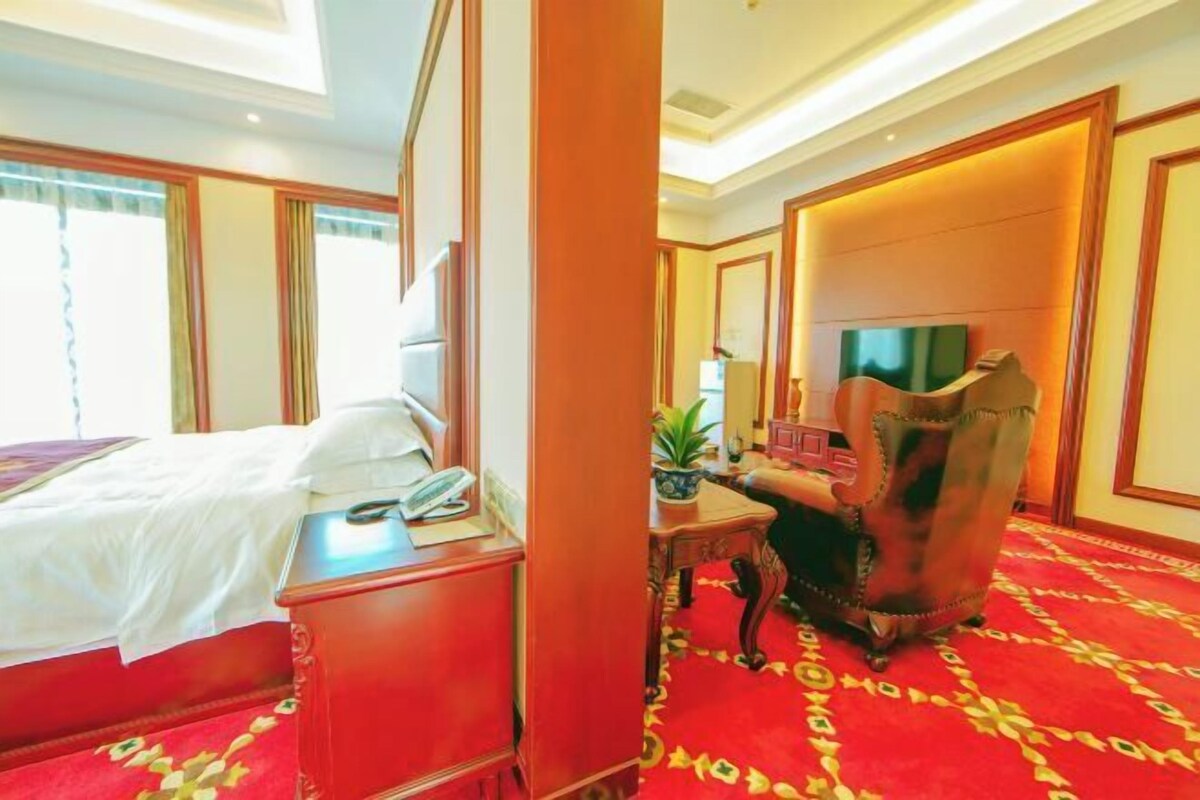 A hotel room with a bed, a desk with a telephone, a chair, a television, and a red carpet.