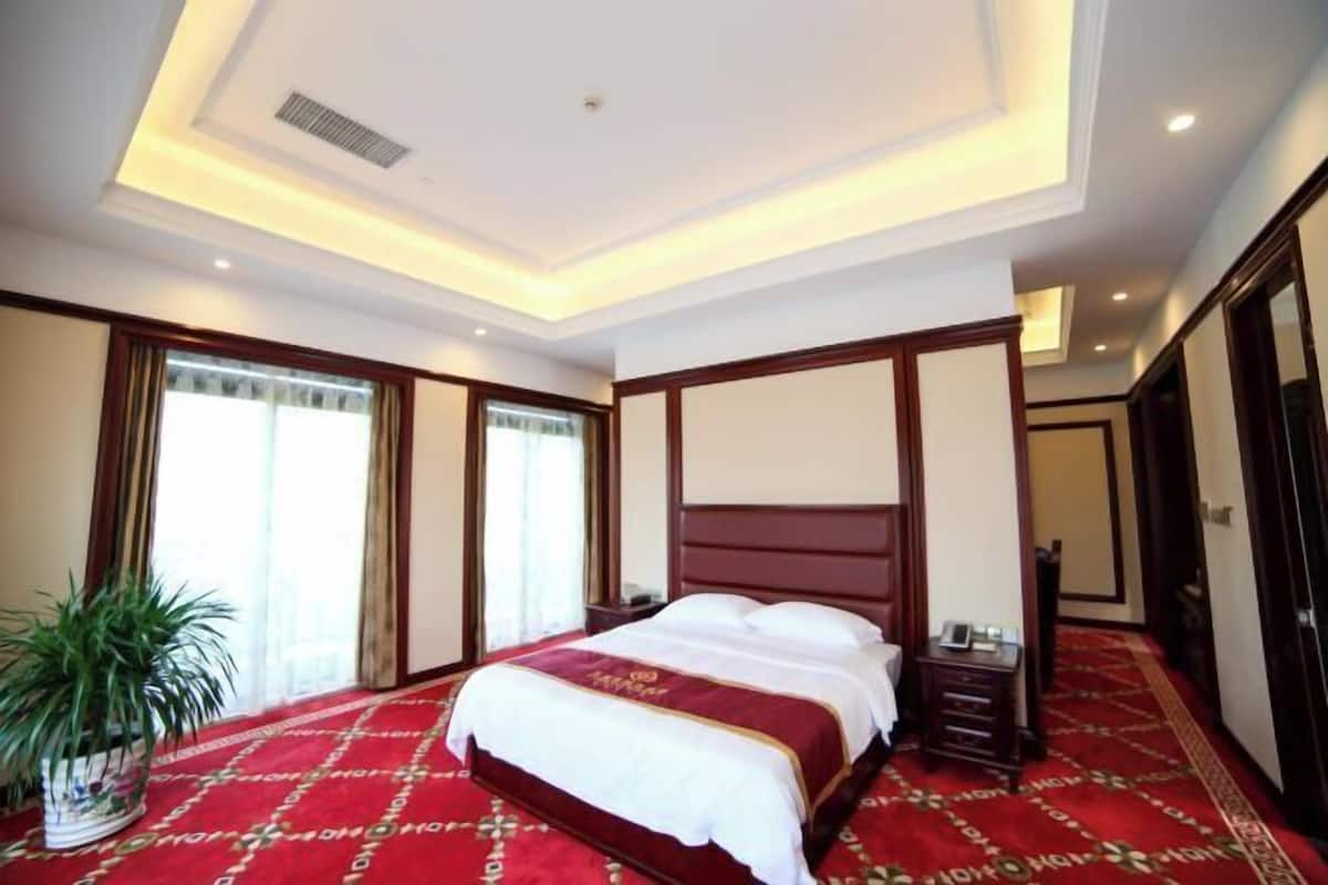 Una habitación de hotel con una cama grande, una mesita de noche, una planta en maceta y una alfombra roja.
