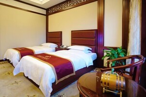 Habitación de hotel con dos camas, cada una con cabecero de madera y ropa de cama blanca. Hay una silla de madera y una mesita con un objeto decorativo.