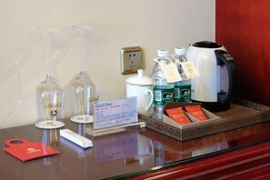 A hotel reception area with a glass display case containing wine glasses, a coffee maker, bottled water, and a kettle. There is a red hotel card and a sign with information.