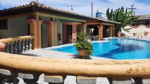 Une piscine dotée d’une terrasse carrelée, d’un espace de repos couvert et d’un bâtiment avec un toit et des colonnes recouverts de carreaux.