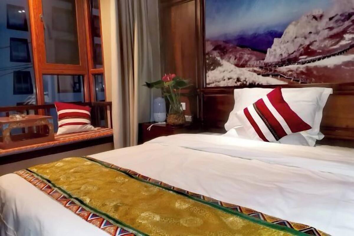 Una camera d'albergo con un letto, una finestra con tende, un quadro raffigurante un paesaggio innevato e un vaso con fiori.