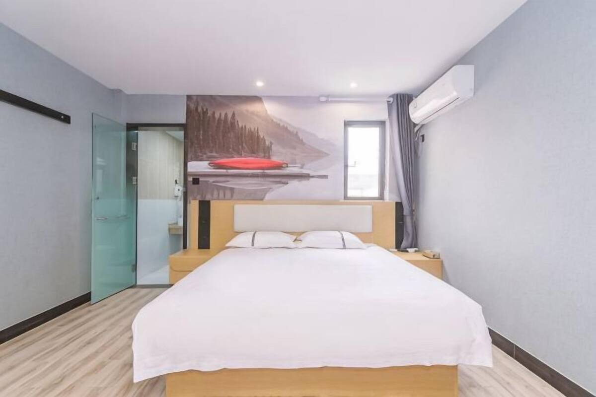 A hotel room with a bed, a painting of a landscape, a window with curtains, and an air conditioning unit.
