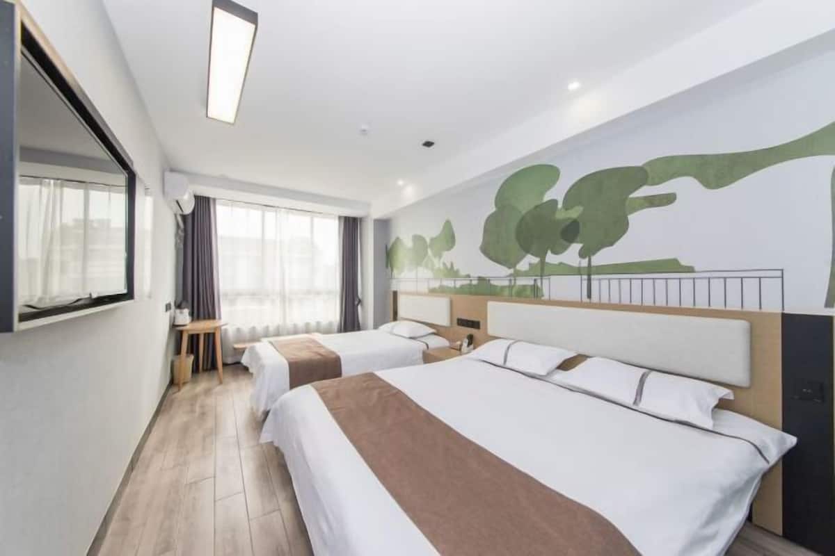 A hotel room with two beds, a large window with curtains, a wall mural, and a small table with a lamp.