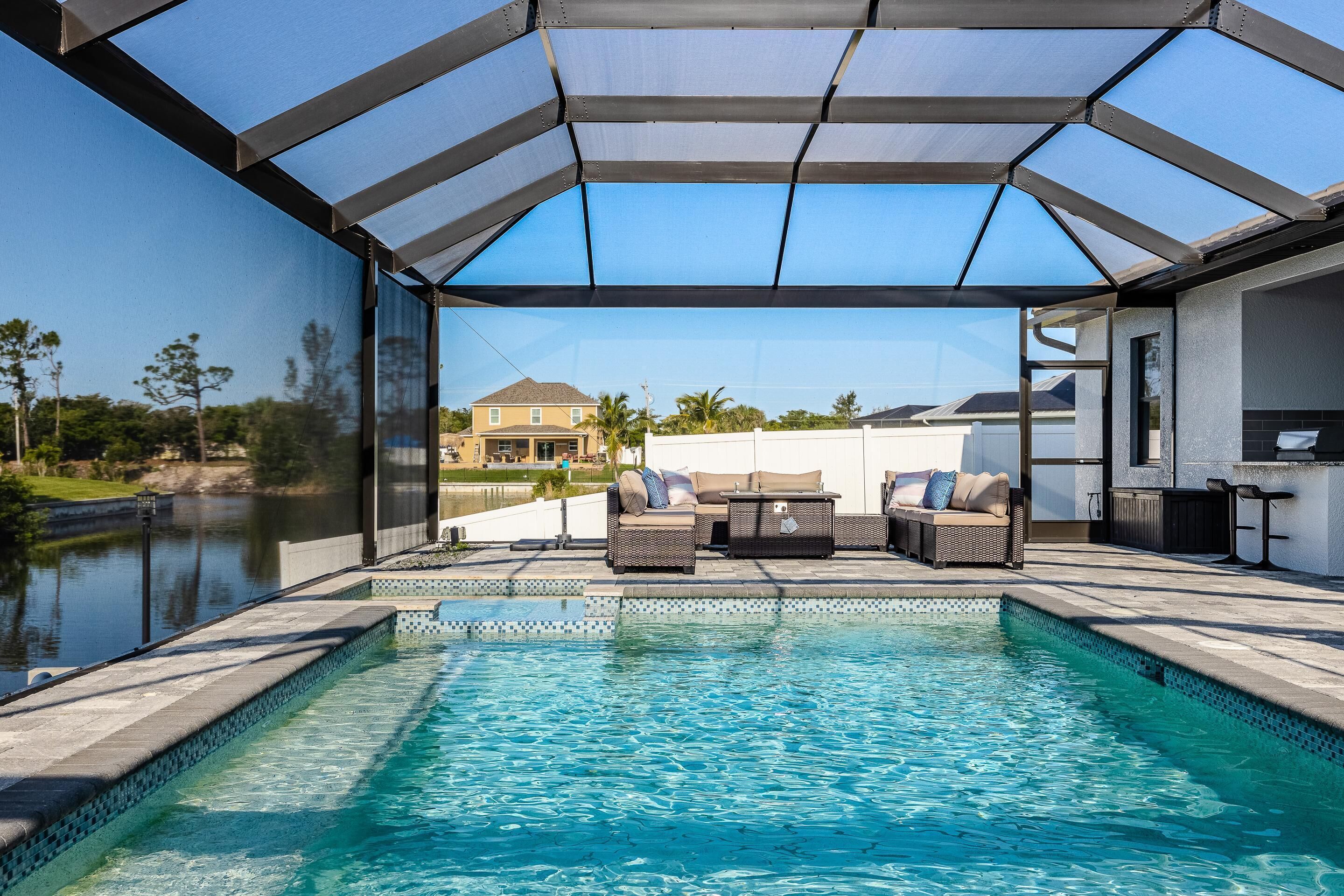 Una piscina con cerramiento de vidrio, mobiliario exterior y vista a una casa al fondo.