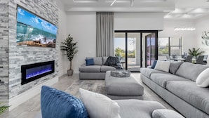 Una sala moderna con pared de piedra, televisor de pantalla plana, chimenea, sofá modular gris y comedor con mesa y sillas.