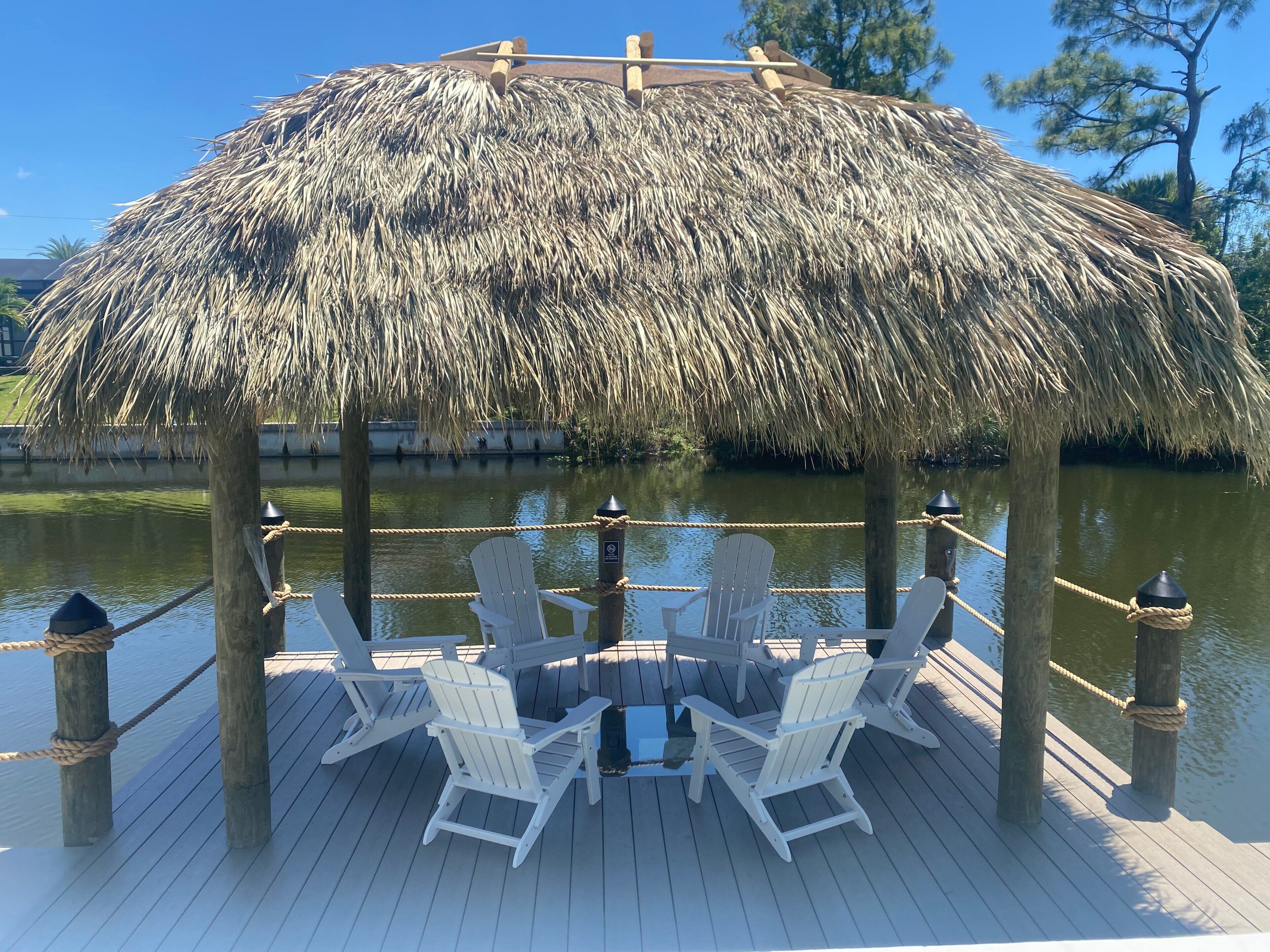 A thatched gazebo with white chairs and a wooden deck over water.