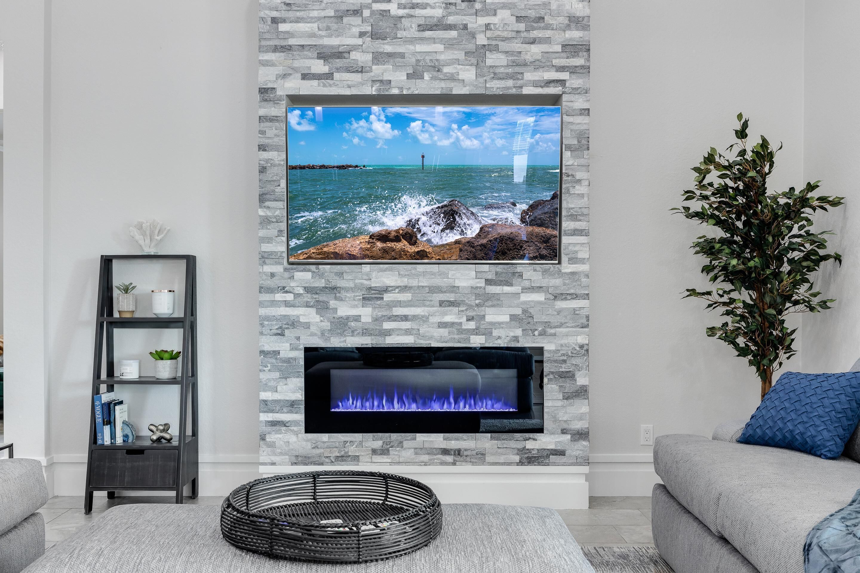 A living room with a stone fireplace, a flat-screen TV mounted above it, a grey sofa, and a side table with a plant.