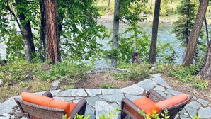 Deux fauteuils en osier avec des coussins orange, placés sur une terrasse en pierre donnant sur une rivière, le tout entouré d’arbres.