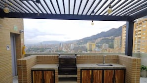 Un área de cocina al aire libre con una parrilla, pileta y armarios de madera, con vista a un paisaje urbano y montañas al fondo.