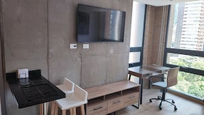 Un espacio de oficina moderno con una televisión de pantalla plana grande montada en una pared de hormigón, un escritorio de madera con una silla y un ventanal con vistas a edificios y vegetación.