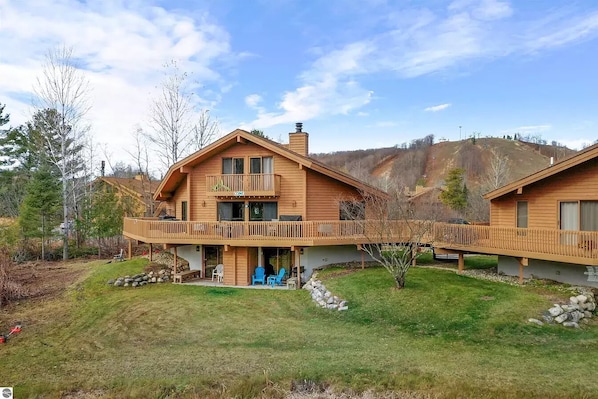 A two-story wooden cabin with a balcony, a stone fireplace, and a view of a hilly landscape.