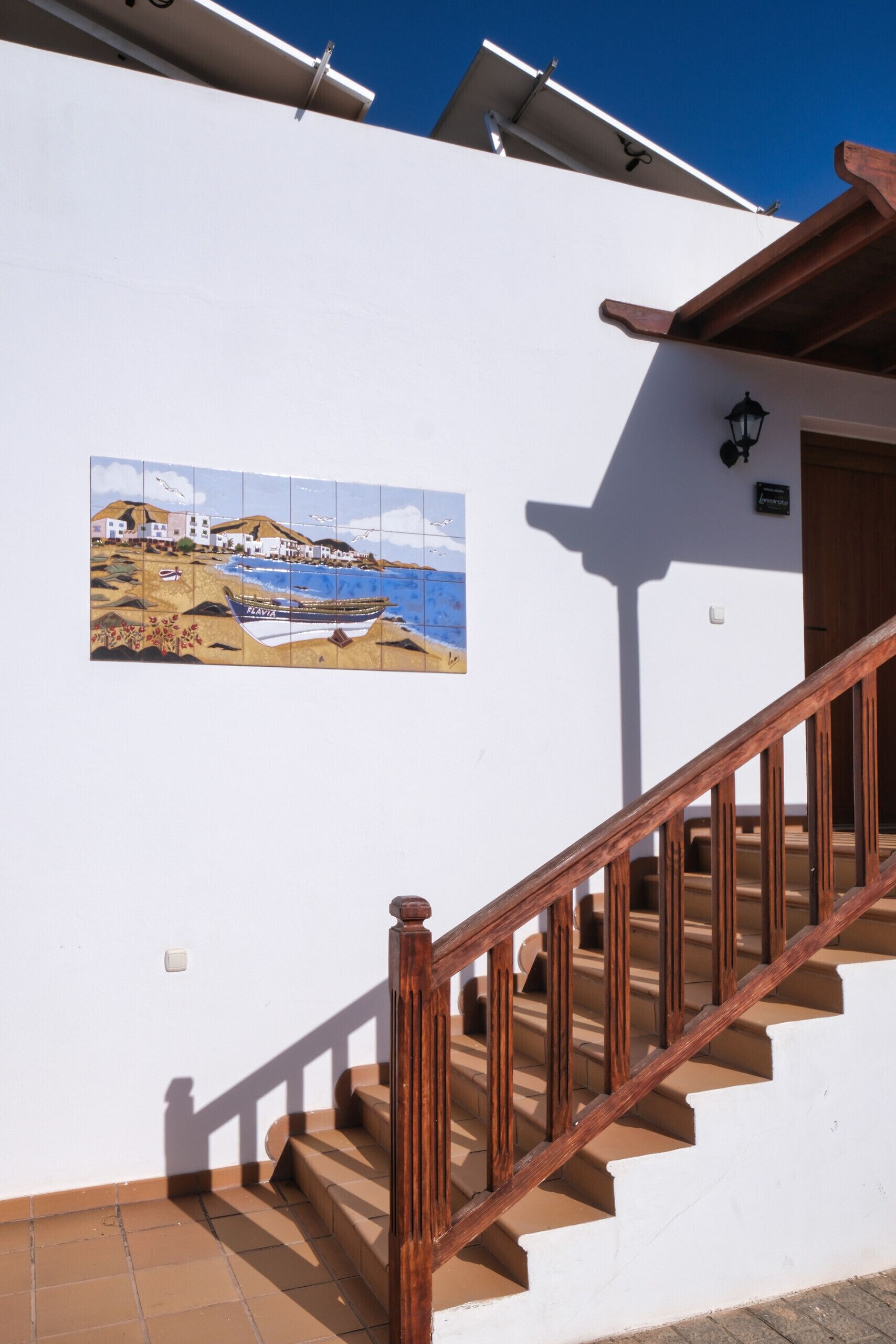 A staircase with wooden railings leading to a door, a tiled wall with a coastal scene, and a clear blue sky.