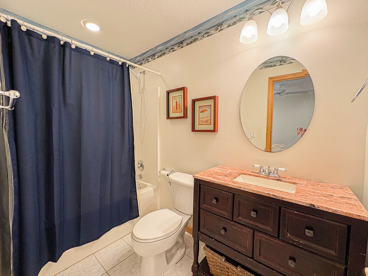 Une salle de bain avec des toilettes blanches, un lavabo doté d’un plan de travail en marbre, un meuble-lavabo en bois foncé, un rideau de douche bleu et un miroir au mur.