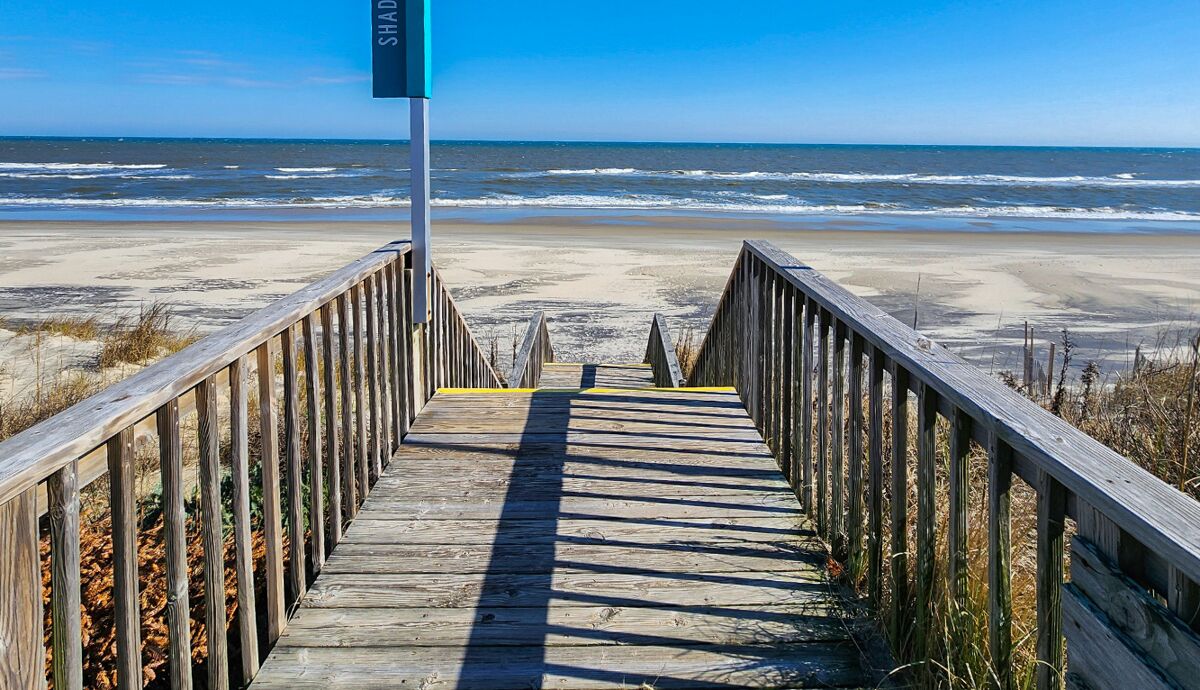 Ein hölzerner Steg führt zu einem Strand, auf dem ein Schild mit der Aufschrift „SHADE“ steht.
