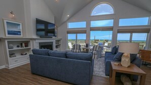 A spacious living room with a large blue sofa, a flat-screen TV, and a fireplace. There is a balcony with outdoor furniture and a view of the ocean. - Baliwest - Gorgeous 5 BR Oceanfront home with a panoramic ocean view! (Southern Shores)