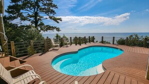 Eine Holzterrasse mit Schwimmbad, umgeben von Stühlen und einem Holzzaun, mit Blick auf ein Gewässer.