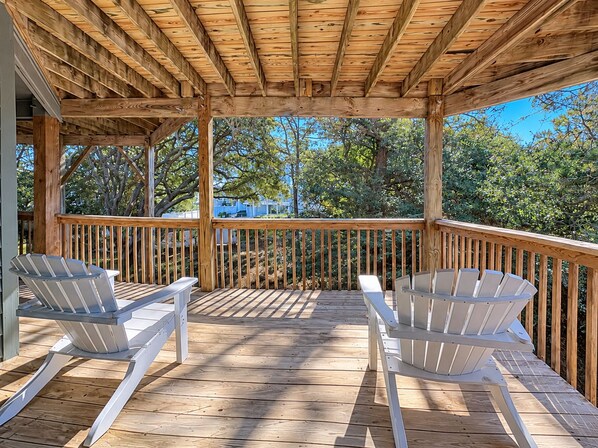 A wooden deck with two Adirondack chairs, a wooden railing, and a view of trees and a blue sky.