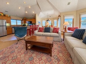 A spacious living area with a sofa, armchair, and coffee table, a kitchen with wooden cabinets and a refrigerator, and large windows providing natural light.