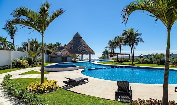 Relax by the pool surrounded by lush greenery and palm trees, with a shaded area and lounge chairs available.