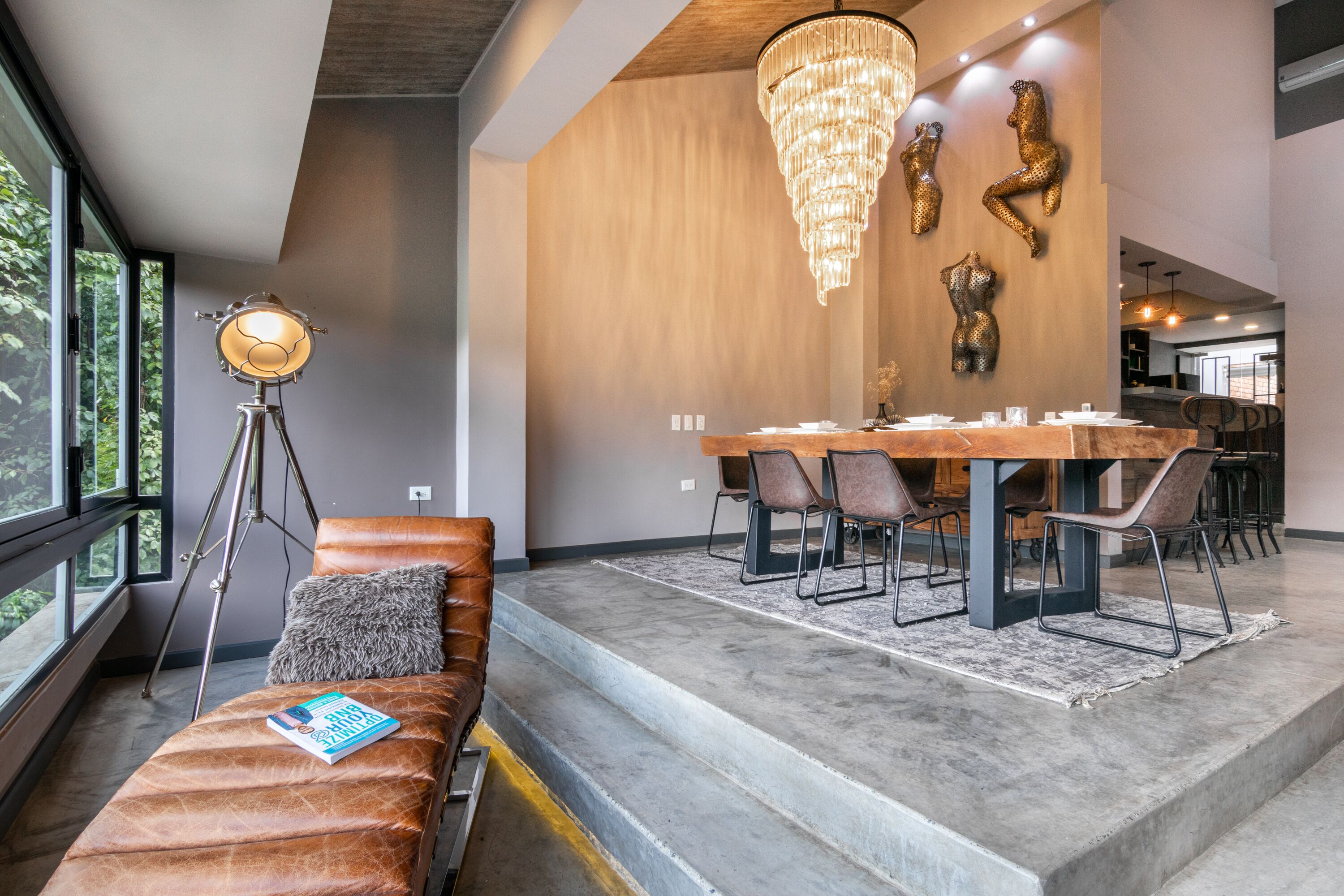 A modern dining area with a wooden table and chairs, a chandelier, and a bench with a book.