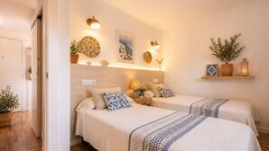 A hotel room with two beds, a wooden headboard, wall-mounted lamps, a shelf with decorative items, and a potted plant. - Codolar – Panoramic Views, Terrace & Parking (Tossa De Mar)
