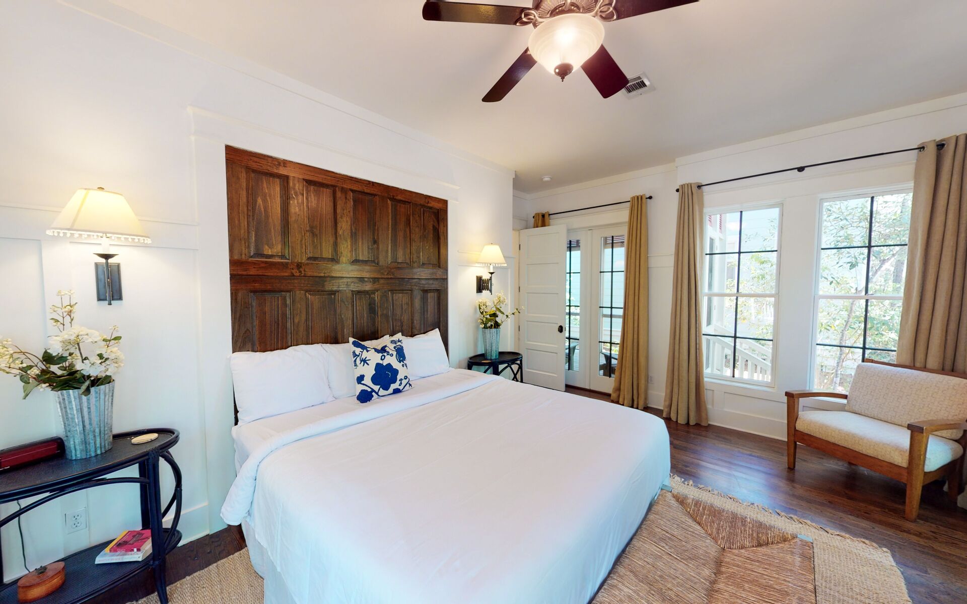 Une chambre d’hôtel avec un grand lit, une tête de lit en bois, un ventilateur de plafond, une fenêtre avec des rideaux et une petite chaise.
