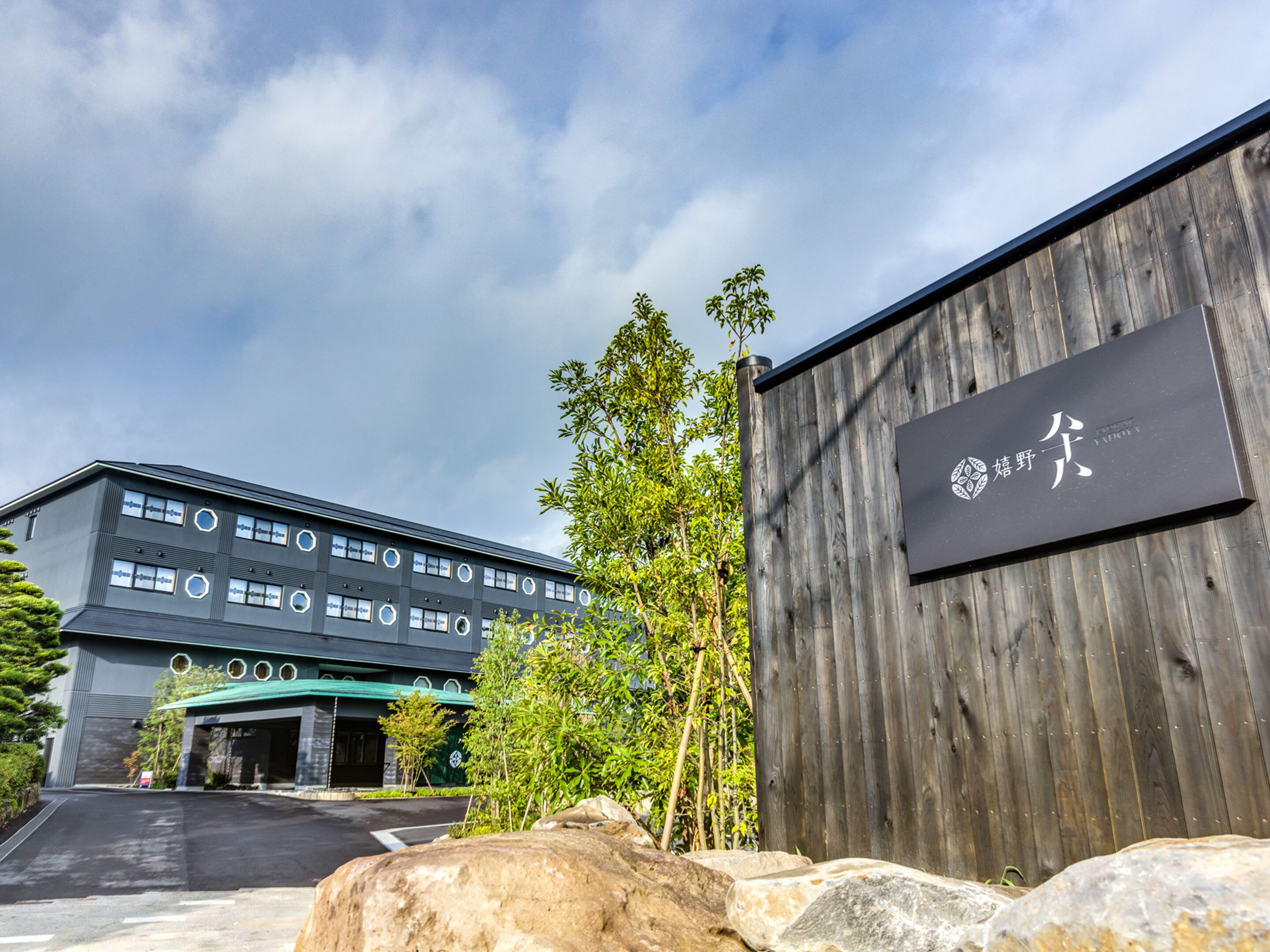 A modern building with a signboard, a parking area, and surrounding greenery.