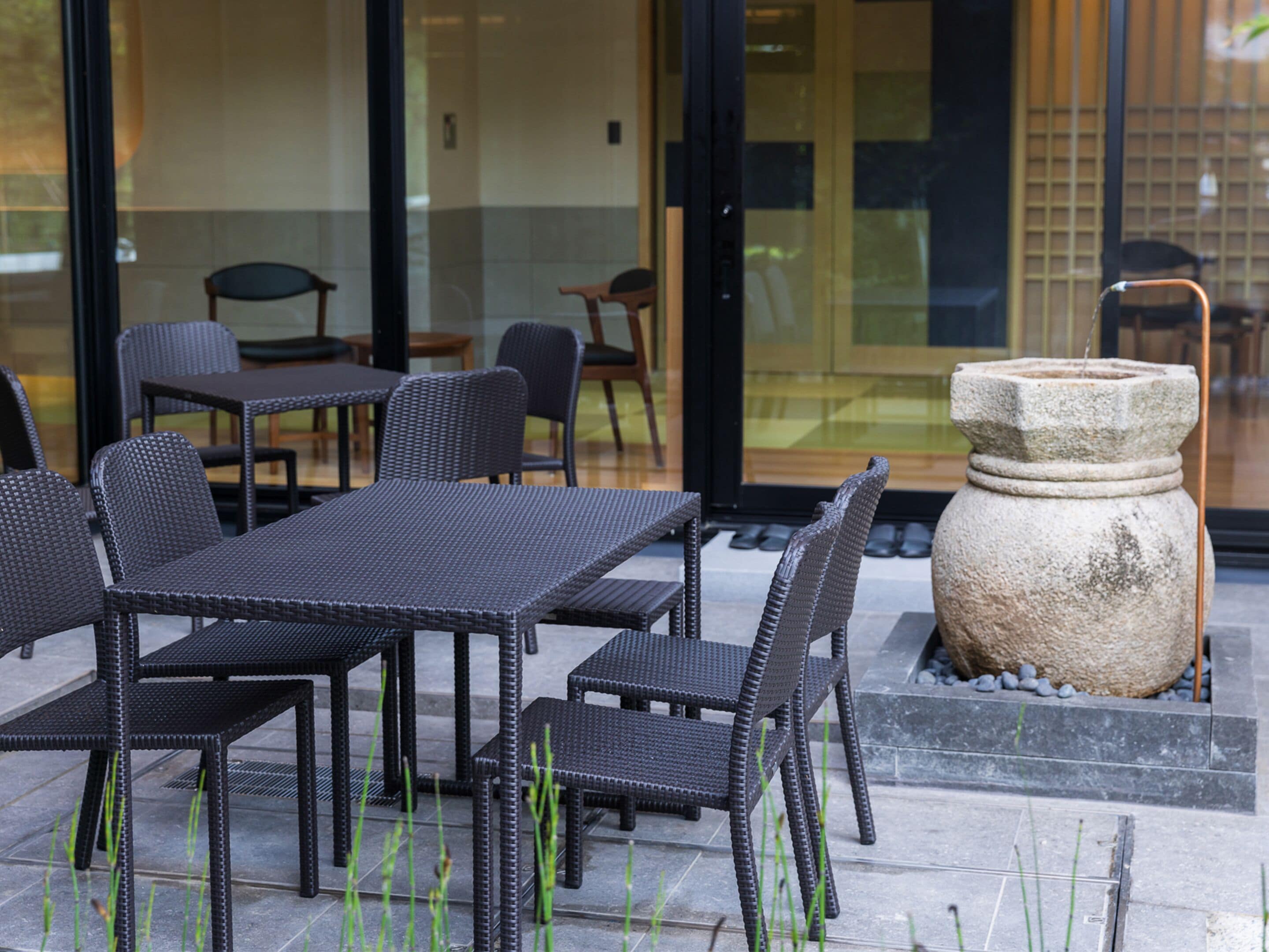 Un espace extérieur aménagé avec des tables et des chaises en osier, un bac à fleurs en pierre et une porte vitrée donnant sur l’intérieur.