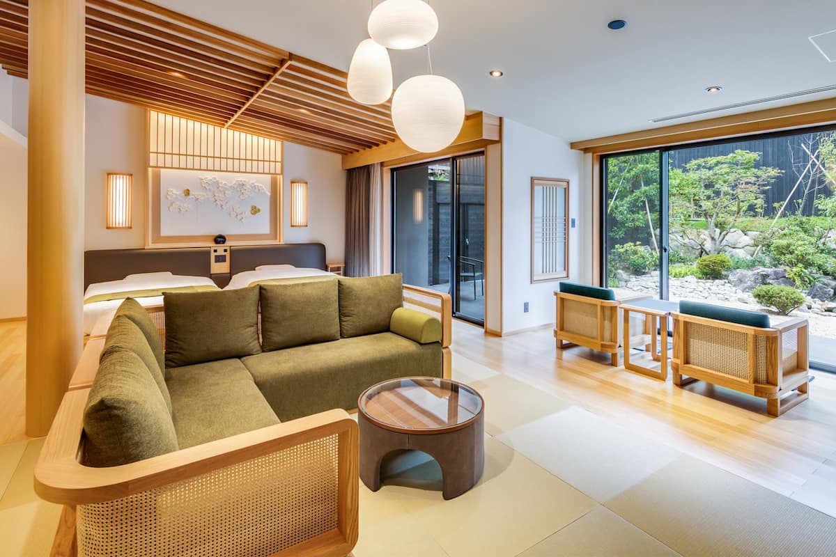A modern hotel room with a bed, a green sofa, a wooden coffee table, and a view of a garden through large windows.