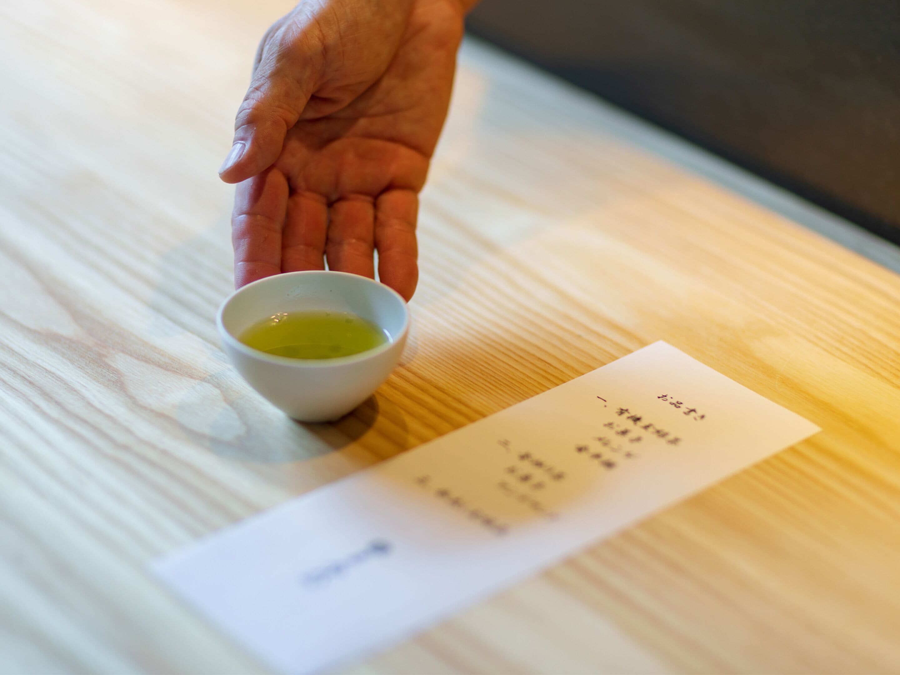Eine Hand hält eine kleine weiße Tasse mit grüner Flüssigkeit, die auf einem Holztisch mit einer Quittung daneben steht.