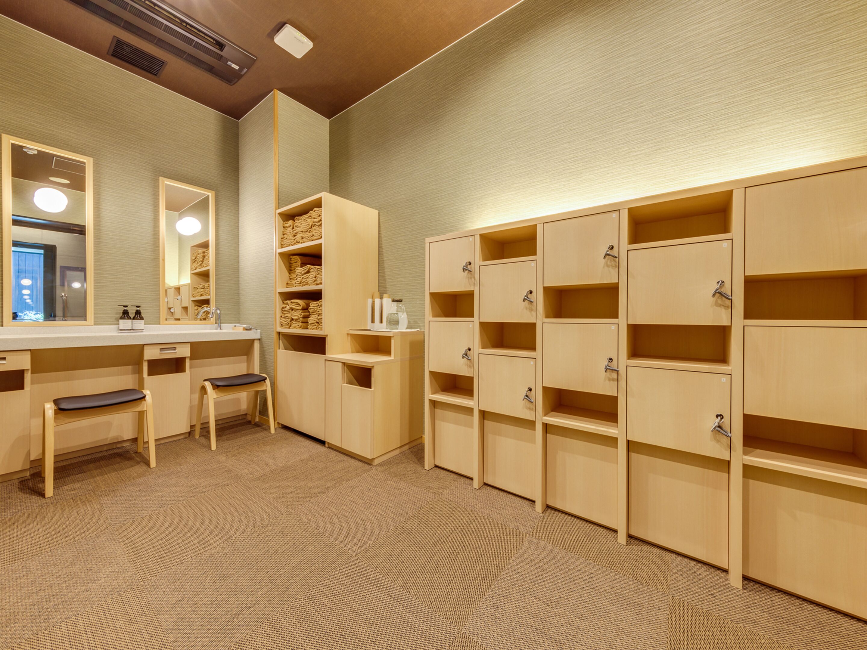 Un baño de hotel con tocador de madera, un banco, repisas con toallas y taquillas.
